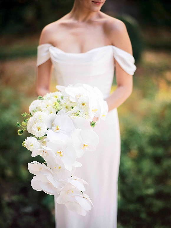 Bridal bouquet of cascading orchids and white garden flowers with greenery, held against an off-shoulder gown in a blurred garden setting