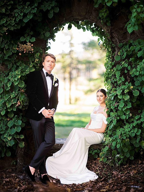 Couple portrait of bride in an off shoulder wedding dress and groom in tuxedo under an ivy archway beside a stone bench in a garden