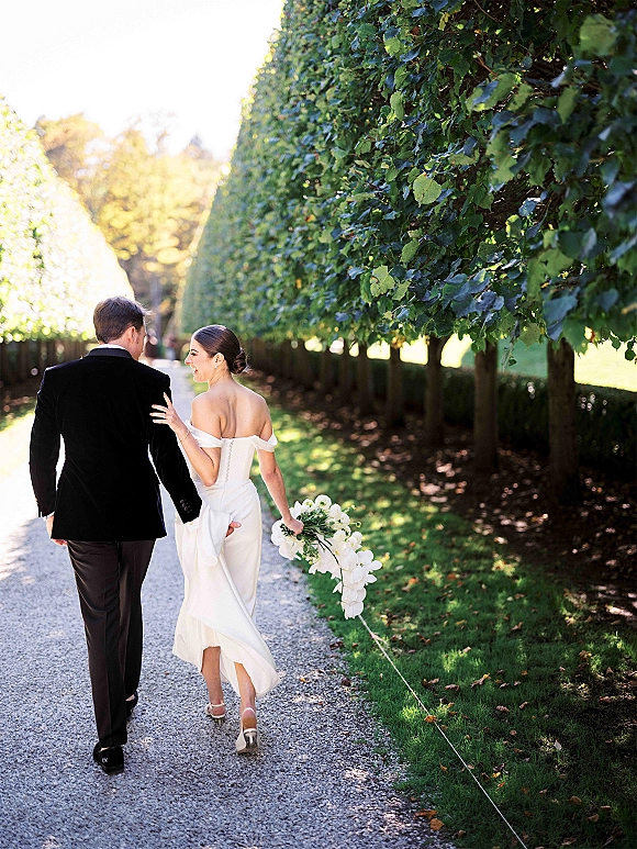Couple portrait of newlyweds walking hand in hand down a sunlit tree-lined path, bride in off-shoulder gown holding bouquet