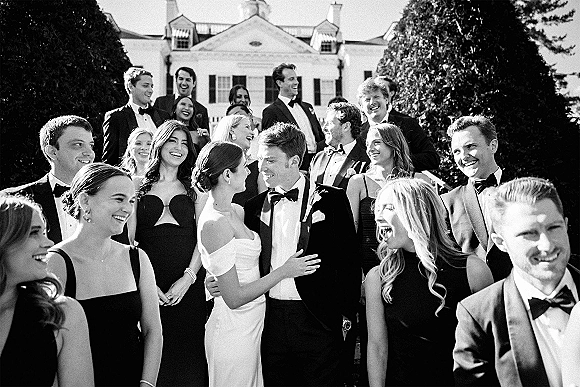 Wedding party portrait of bride and groom with friends in tuxedos and black dresses, laughing on mansion stairs with trees behind