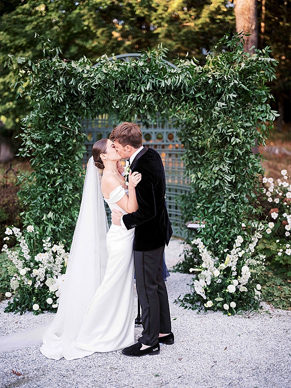 Wedding kiss portrait of bride and groom kissing under a greenery arch, her cathedral veil flowing beside white florals on a gravel path