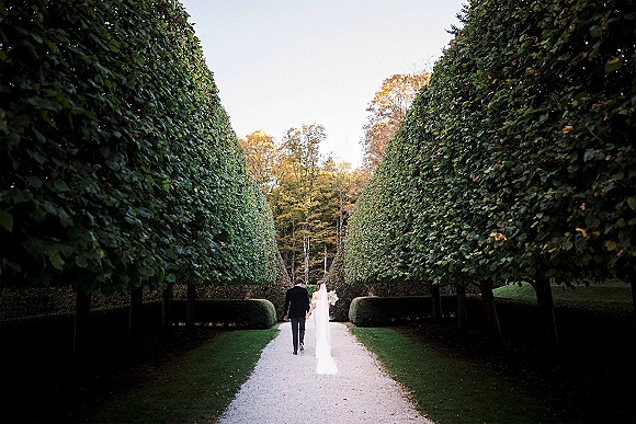 Couple walking away hand in hand, bride and groom walking down a tree-lined garden path with veil, bouquet, and tuxedo in fall light