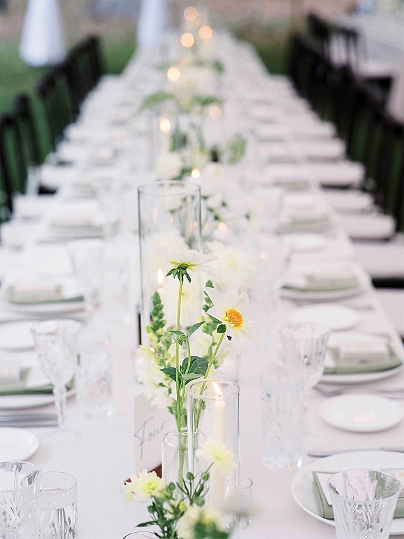 Reception tablescape on a long banquet table with white linens, glass cylinder vases, taper candles, white blooms, and greenery outdoors