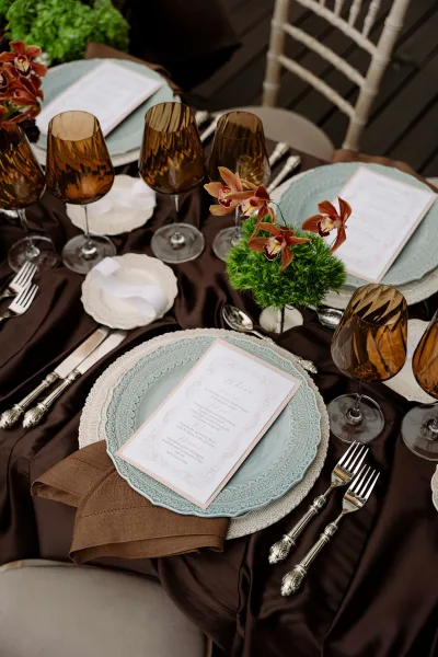 Reception tablescape with wedding place setting, amber wine glasses, blue plates, lace chargers, printed menu cards, and orchid centerpiece indoors