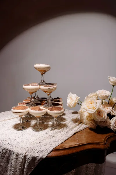 Champagne tower of coupe glasses with foam-topped cocktails on a lace tablecloth, styled with white roses before an arched niche