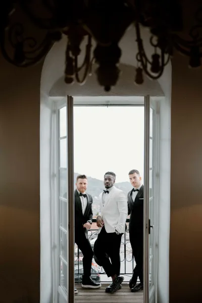Groomsmen portrait of the groom with groomsmen in tuxedos, one in a white jacket, leaning by open French doors on a balcony above a hillside town