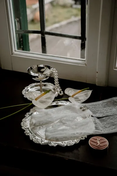 Bridal accessories flatlay with pearl necklace, lace gloves, earrings, and calla lilies arranged on a silver tray by window light