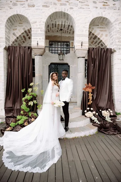 Couple portrait of bride in veil and long train holding a calla lily bouquet beside groom in white tuxedo under a stone archway