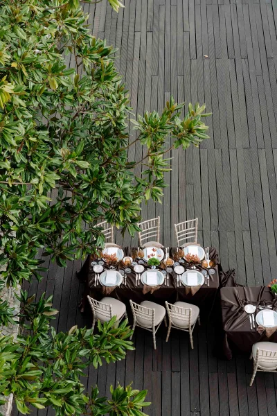 Reception tablescape with a brown tablecloth, white Chiavari chairs, amber glassware, menus, candles, and floral greenery on a wood deck under trees