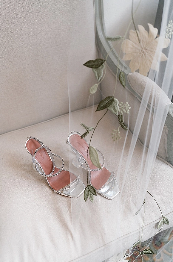 Bridal shoes with wedding shoe detail, rhinestone strap clear heels paired with a floral embroidered veil on an upholstered chair