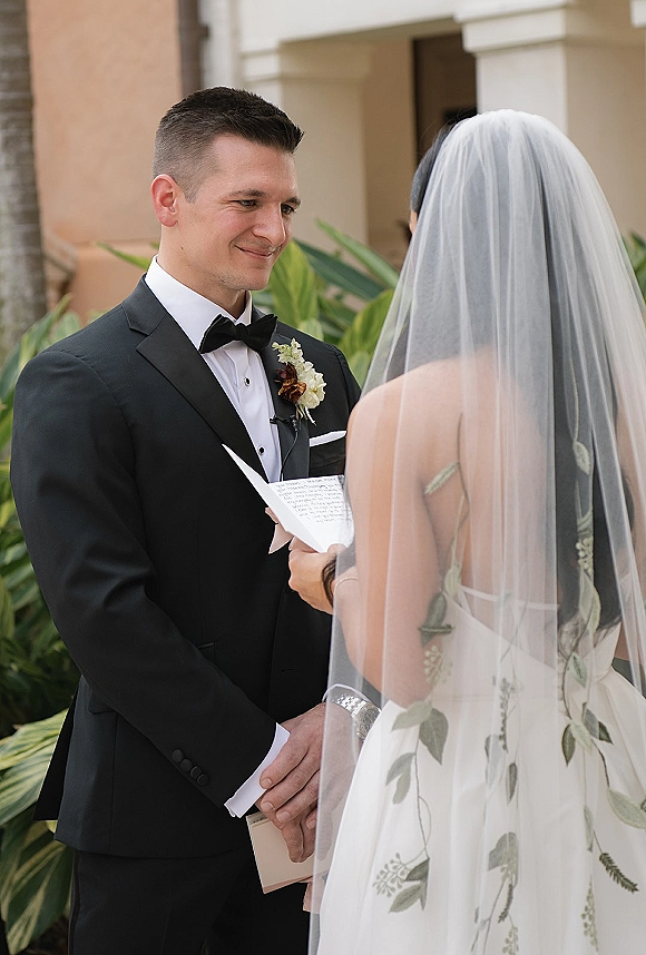 Wedding vows as the bride reading vows in a veil and gown faces a smiling tuxedoed groom with boutonniere, framed by tropical greenery and columns