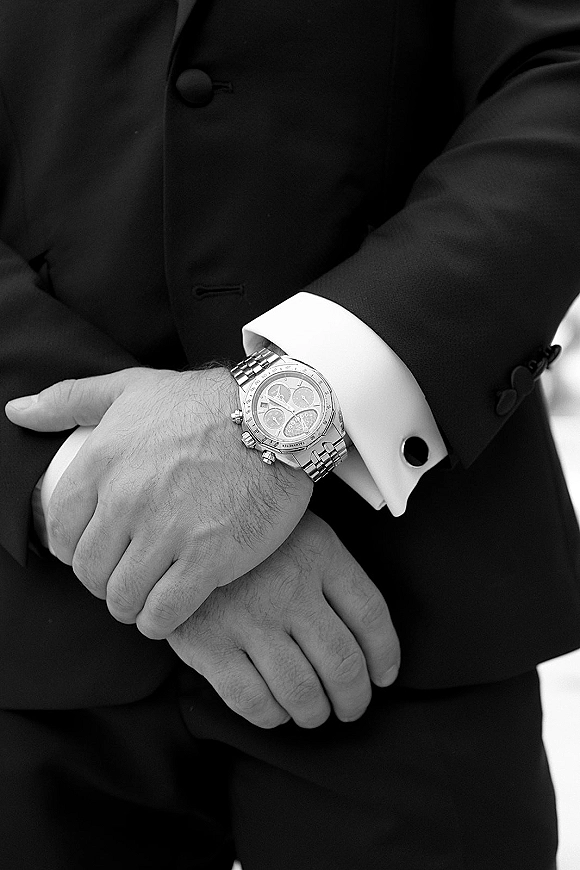 Groom watch detail showing a silver chronograph wristwatch beside a cufflink on a white French cuff under a black tuxedo sleeve