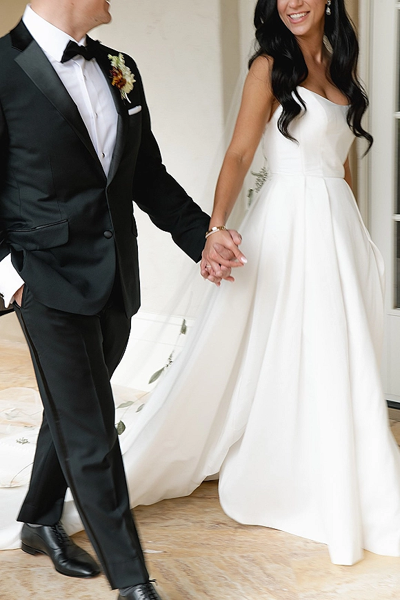 Couple portrait of bride and groom holding hands, her ball gown and embroidered cathedral veil trailing beside his black tux by a doorway
