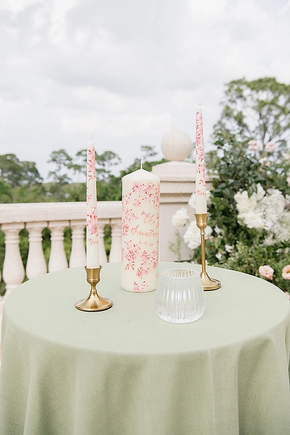 Wedding table candles with hand painted wedding candles on brass candlesticks atop a green tablecloth, set by a stone balustrade garden backdrop