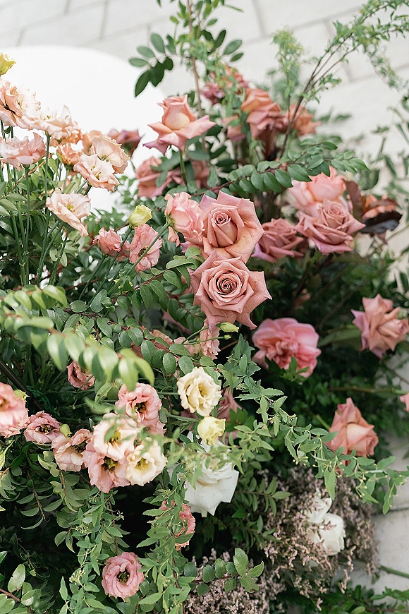 Wedding florals with a blush rose arrangement, peach and blush roses, spray roses, lisianthus, and greenery against a white brick wall
