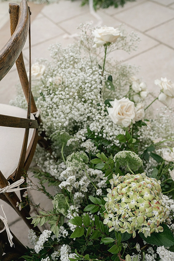 Aisle florals and wedding aisle flowers with white roses, hydrangea, and greenery beside a wooden chair on stone tile floor