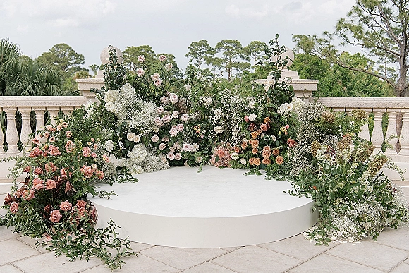 Wedding ceremony backdrop with blush and white rose arrangements and greenery garland on a circular platform on a stone terrace patio
