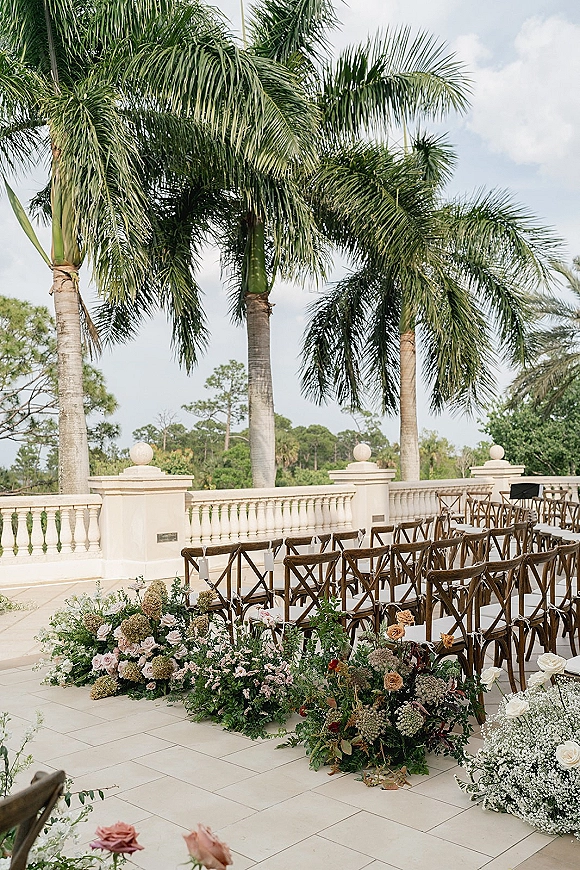 Outdoor ceremony setup with garden ceremony aisle, crossback chairs, and white rose meadow florals on a stone terrace with palm trees under blue sky