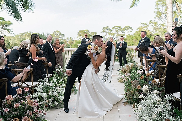 Ceremony kiss as groom dips bride in strapless dress, bouquet and veil flowing beside rose petal aisle on palm-lined terrace
