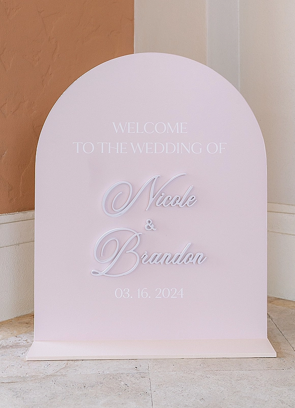 Wedding welcome sign with an arched board and raised white lettering in serif and script, set against a stucco wall and stone tile floor