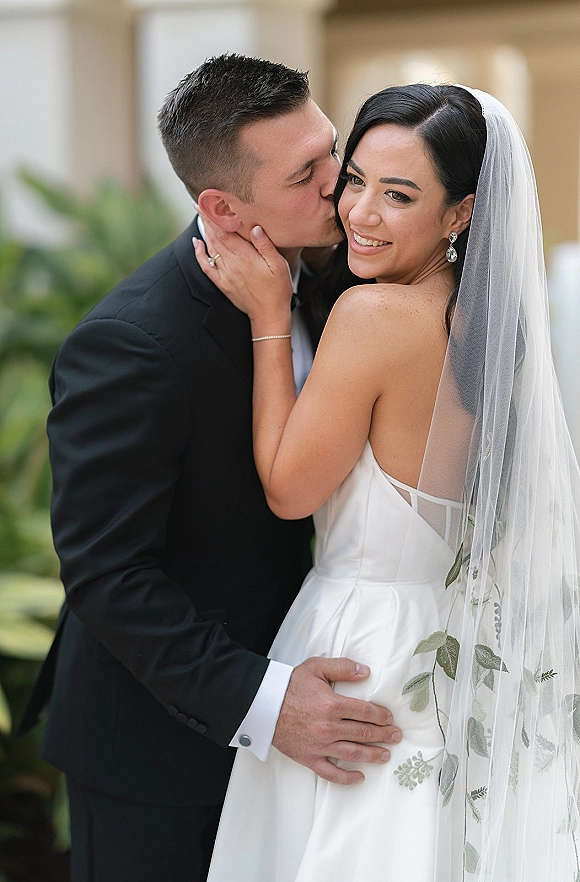 Wedding couple portrait with groom kissing bride’s cheek as she looks over her shoulder, veil and strapless dress in outdoor greenery backdrop