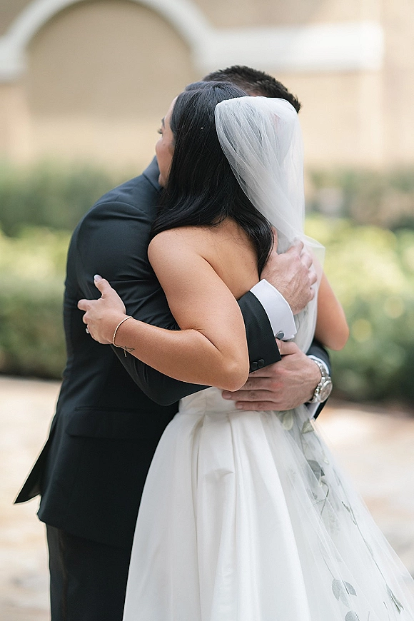 Wedding hug as bride and groom embrace tightly from behind, her veil over shoulder, in an outdoor courtyard with arches and greenery