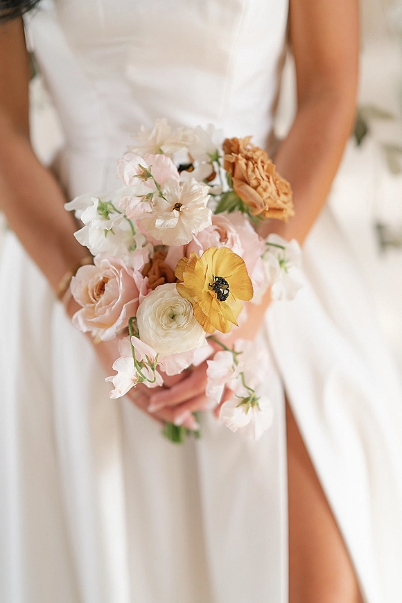 Bridal bouquet, colorful bridal bouquet of roses, ranunculus, and sweet peas with greenery held against a simple wedding dress indoors