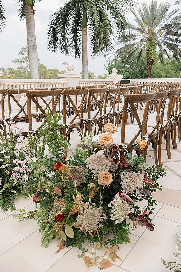 Ceremony seating with cross back ceremony chairs tied in white ribbons, lining an aisle with blush florals on a stone terrace by palms