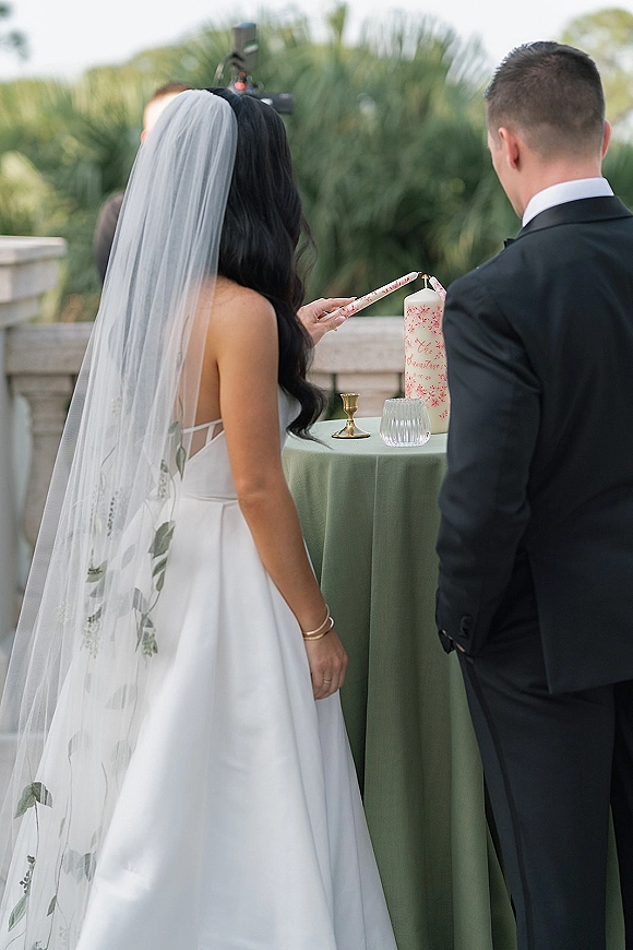 Unity candle ceremony as bride in veil and groom in tux light taper candles beside a pillar candle on a round table with greenery behind