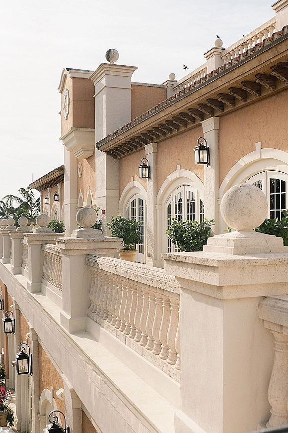 Wedding venue exterior with a stone balustrade, arched windows, and wall lanterns on a stucco facade with palms and tiled roof under blue sky