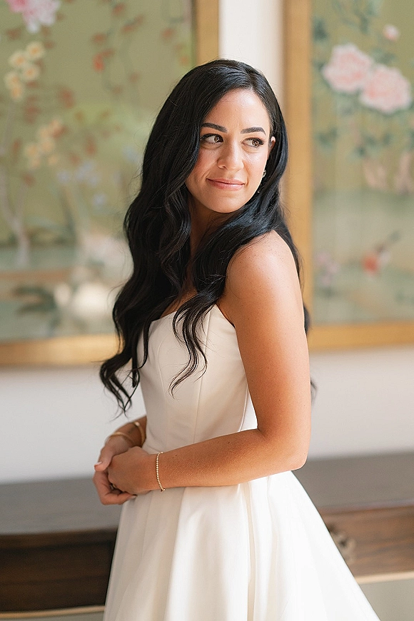 Bridal portrait of a bride in a strapless wedding dress looking over her shoulder, long wavy hair and bracelet before floral wall art indoors