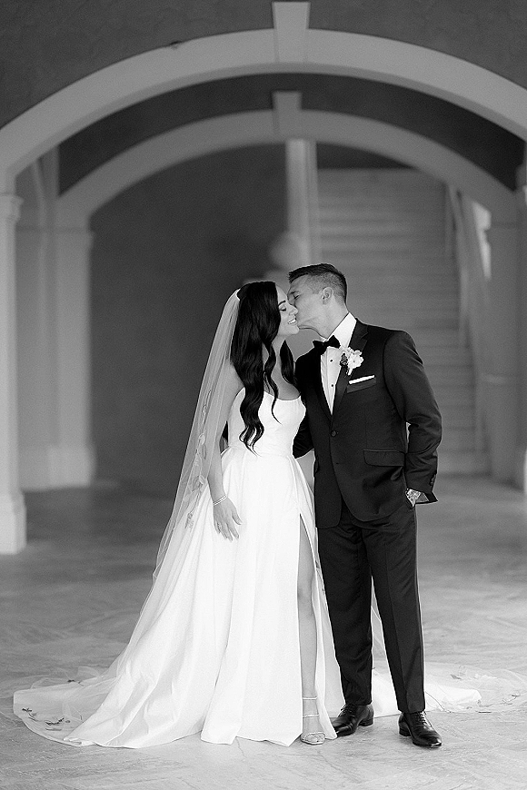 Wedding kiss portrait of bride and groom kissing in a black and white wedding portrait under arched columns, her veil flowing behind them