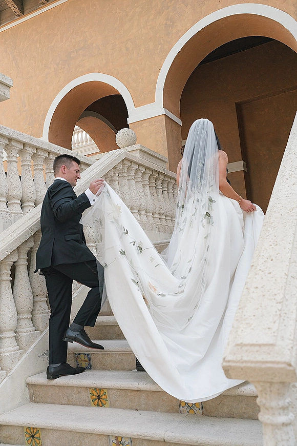 Couple portrait of groom carrying dress train as bride walks upstairs in a cathedral veil, framed by stone staircase and archways