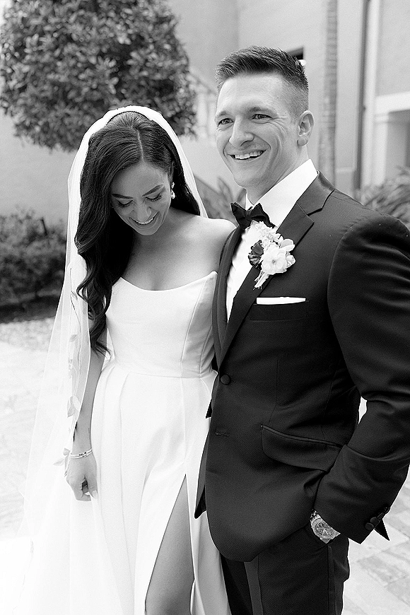 Couple portrait in black and white wedding portrait style, bride in strapless thigh-slit gown and veil walks close with smiling groom in tuxedo courtyard