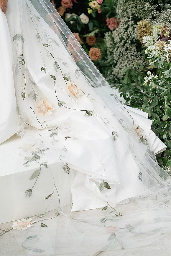 Bridal veil close-up with floral embroidered veil draped over the wedding dress skirt, sheer tulle train against soft greenery backdrop