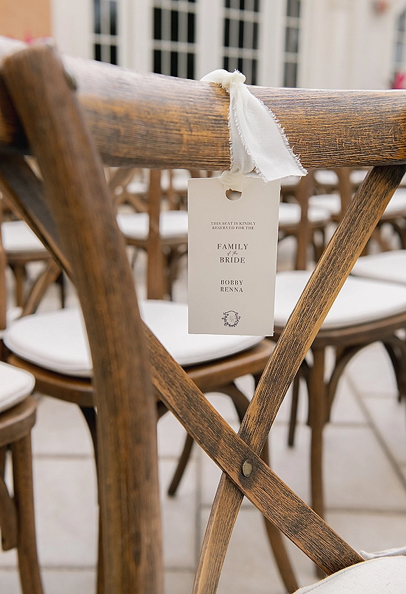 Wedding reserved seat sign tied with a white ribbon on a wooden crossback chair with cushion amid ceremony chairs on tiled floor by windows