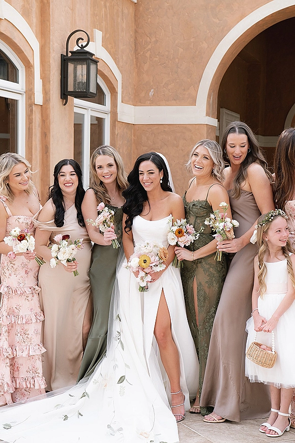 Bridesmaid group photo with bride and bridesmaids laughing, holding bouquets by a stucco wall and arched doorway, veil flowing