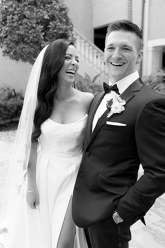 Wedding couple portrait, bride and groom laughing together in a courtyard by stairs, her long veil and strapless dress beside his tuxedo