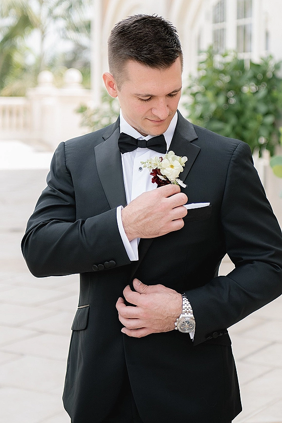 Groom portrait in a black tuxedo adjusting his boutonniere under white arches, with columns, greenery, and palm trees behind him