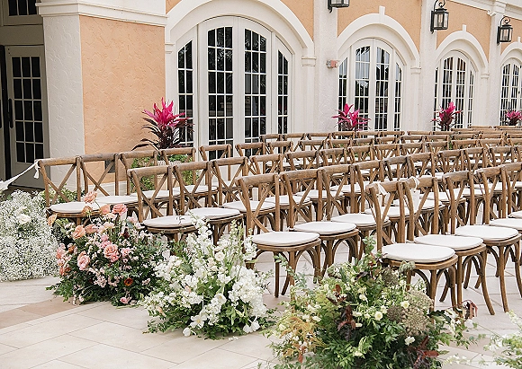Ceremony seating with cross back chair wedding rows and white cushions, lined by blush rose aisle florals in a stucco courtyard with arched windows