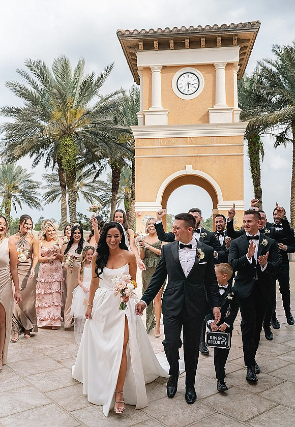 Wedding recessional as bride and groom walking hand in hand past cheering party under a clock tower arch with palm trees and bouquet