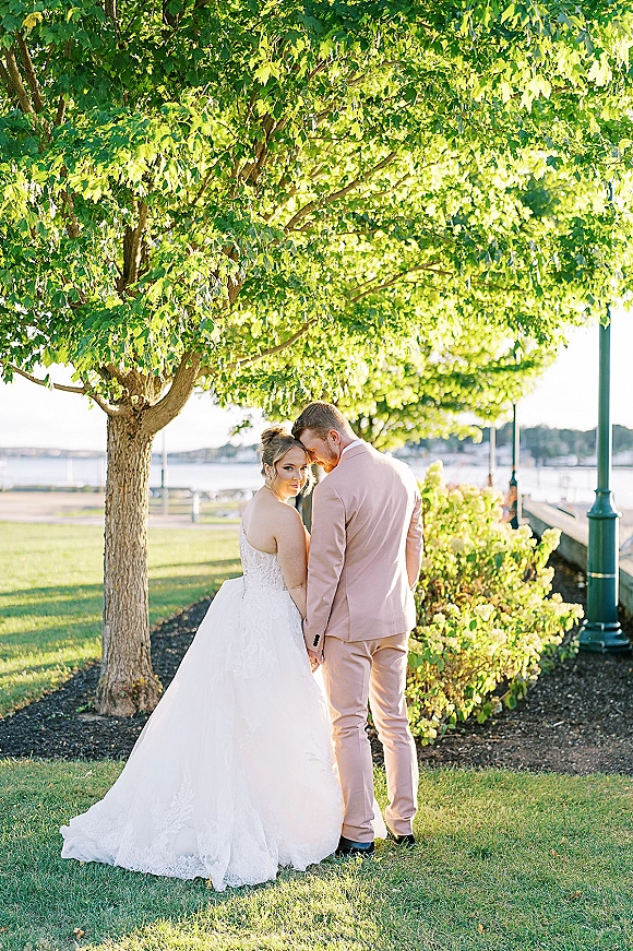 Couple portrait of bride and groom holding hands, bride in lace gown and bridal updo, groom in blush suit on a waterfront lawn path