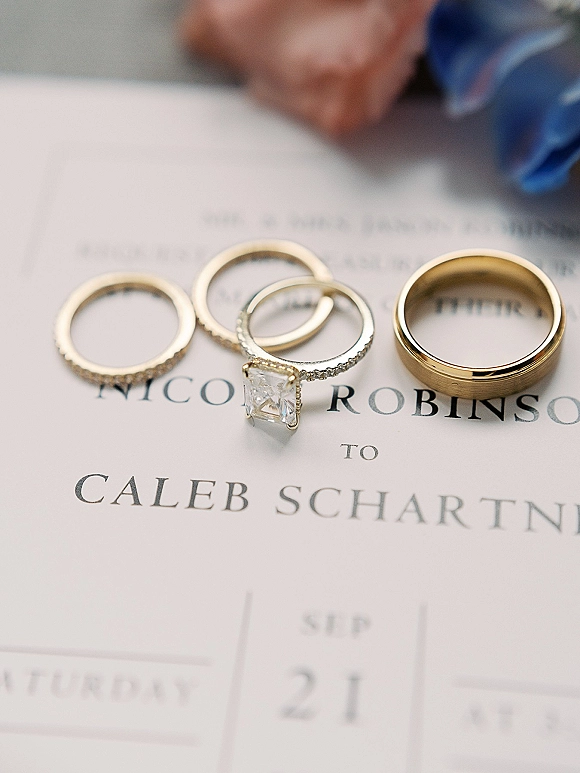 Wedding rings and engagement ring close up featuring a diamond set beside a gold wedding band on a wedding invitation with blurred hands