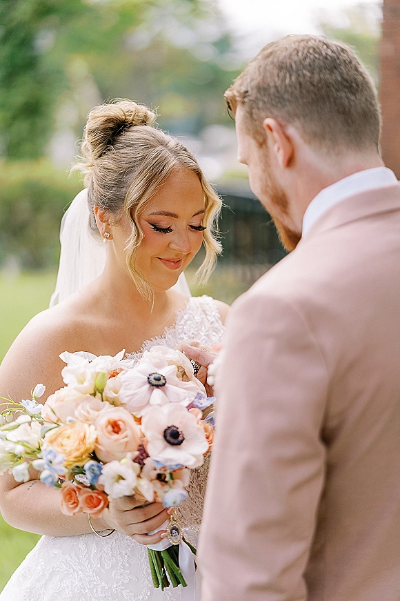 First look moment outdoors as bride holds bouquet and looks down, lace wedding dress and veil beside groom in blush suit by brick wall