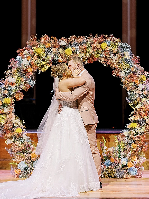 Wedding kiss as bride in veil and lace gown embraces groom in tan suit under circular floral arch on a wooden stage with dark backdrop
