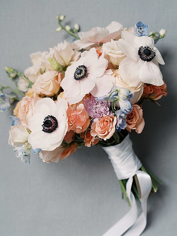 Bridal bouquet with white anemones, peach roses and delphinium, tied with a white ribbon wrap on a gray surface flat lay