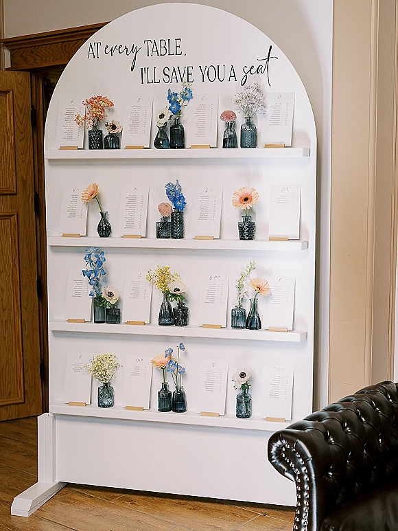 Seating chart display with a round wedding seating chart sign in calligraphy, escort cards on white shelves, with blue glass bud vases indoors