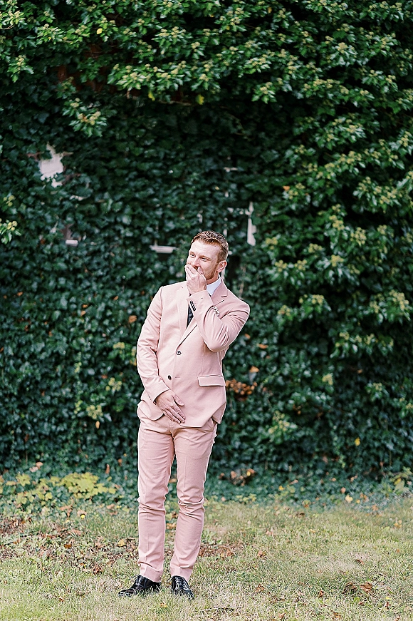 Groom portrait in a blush groom suit with black tie, covering his mouth while laughing beside an ivy wall on a green lawn outdoors