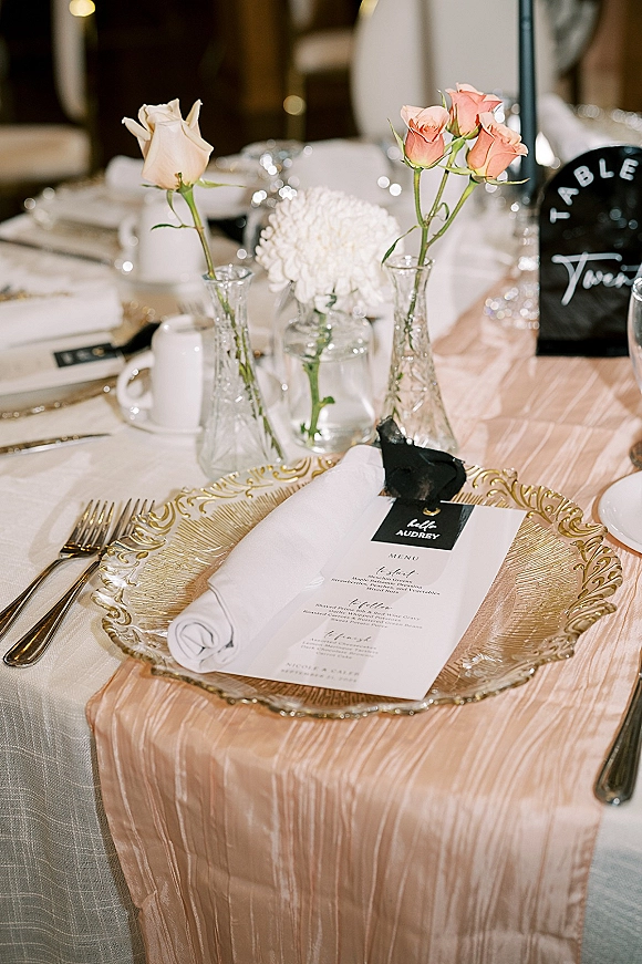 Reception tablescape wedding place setting with gold charger plate, white napkin, menu card, black ribbon name tag, and peach roses in bud vases