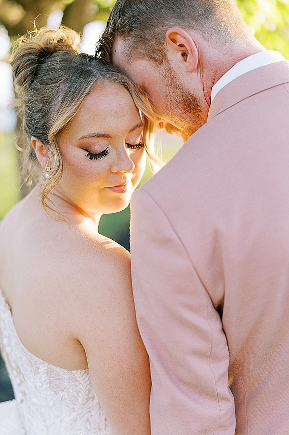 Couple portrait of bride and groom embrace, forehead to forehead in sunlight, her lace bodice and pearl studs framed by trees